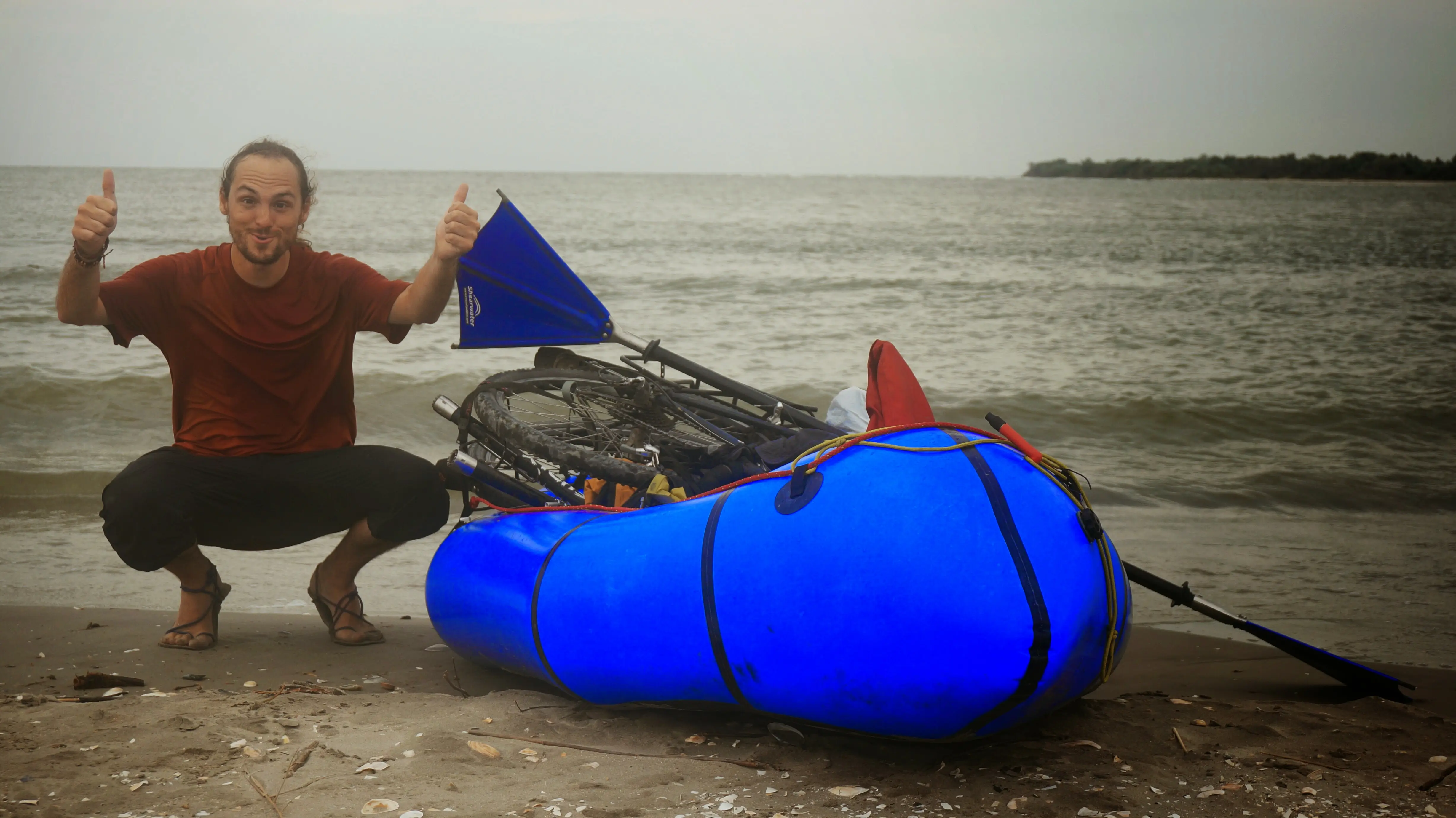A man giving thumbs up next to a blue inflatable raft with a bicycle strapped on top at a beach.